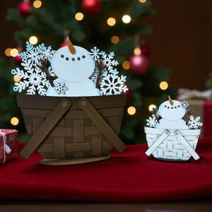 May include: Two wooden snowman decorations in baskets. The snowmen have carrot noses, stick arms, and snowflake accents. The baskets are brown and white, and the background includes a Christmas tree and red tablecloth.