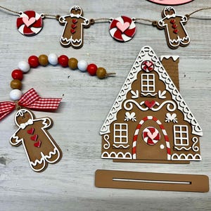 K&ouml;nnte beinhalten: Ein Lebkuchenhaus-Anh&auml;nger mit wei&szlig;en Zuckergussdetails, roten Bonbonakzenten und einer braunen Basis. Das Haus ist mit roten und wei&szlig;en Zuckerstangen und Herzen verziert. Der Anh&auml;nger ist Teil einer Lebkuchen-Girlande mit Lebkuchenm&auml;nnern und Zuckerstangen-Anh&auml;ngern.