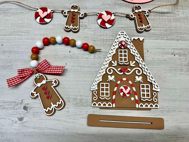 K&ouml;nnte beinhalten: Ein Lebkuchenhaus-Anh&auml;nger mit wei&szlig;en Zuckergussdetails, roten Bonbonakzenten und einer braunen Basis. Das Haus ist mit roten und wei&szlig;en Zuckerstangen und Herzen verziert. Der Anh&auml;nger ist Teil einer Lebkuchen-Girlande mit Lebkuchenm&auml;nnern und Zuckerstangen-Anh&auml;ngern.