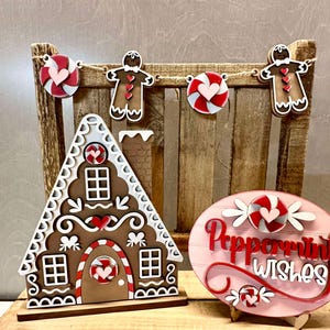 K&ouml;nnte beinhalten: Weihnachtsdekoration aus Holz mit einem Lebkuchenhaus, einem Schild in Form eines Pfefferminzbonbons und einer Girlande mit Zuckerstangen und Lebkuchenm&auml;nnern. Das Schild lautet "Peppermint Wishes".