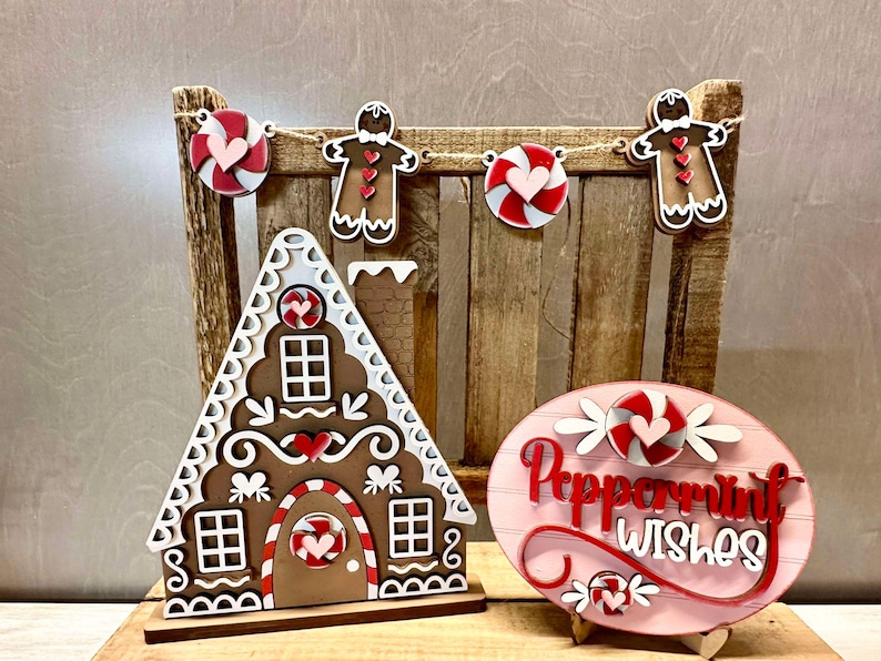 K&ouml;nnte beinhalten: Weihnachtsdekoration aus Holz mit einem Lebkuchenhaus, einem Schild in Form eines Pfefferminzbonbons und einer Girlande mit Zuckerstangen und Lebkuchenm&auml;nnern. Das Schild lautet "Peppermint Wishes".