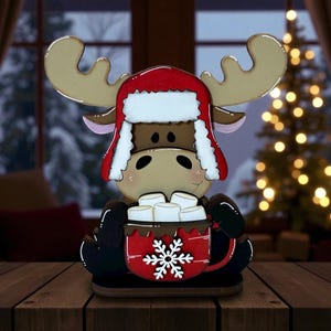 May include: A wooden moose figurine wearing a red and white winter hat. The moose is holding a red mug of hot chocolate topped with marshmallows and a white snowflake design. The figurine is on a wooden base.