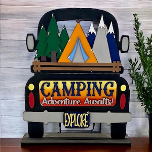 May include: A black wooden truck with a camper tent, trees, and mountains on the back. The truck has a sign that says "Camping Adventure Awaits!" and a license plate that says "Explore".