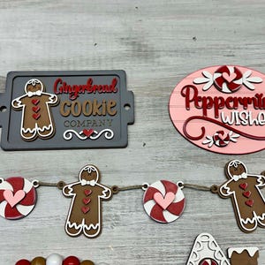 K&ouml;nnte beinhalten: Ein Schild aus Holz f&uuml;r eine Lebkuchen-Kekse-Firma mit einem Lebkuchenmann und dem Text "Gingerbread Cookie Company". Ein Schild aus Holz mit Pfefferminz-W&uuml;nschen mit einem Zuckerstangen und dem Text "Peppermint Wishes". Eine Girlande aus Holz mit Lebkuchenm&auml;nnern und Zuckerstangen.