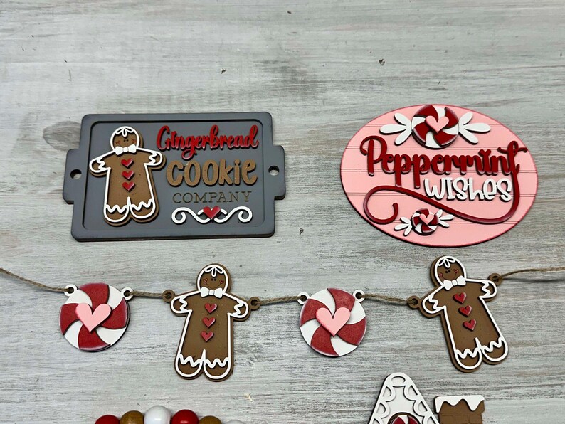 K&ouml;nnte beinhalten: Ein Schild aus Holz f&uuml;r eine Lebkuchen-Kekse-Firma mit einem Lebkuchenmann und dem Text "Gingerbread Cookie Company". Ein Schild aus Holz mit Pfefferminz-W&uuml;nschen mit einem Zuckerstangen und dem Text "Peppermint Wishes". Eine Girlande aus Holz mit Lebkuchenm&auml;nnern und Zuckerstangen.