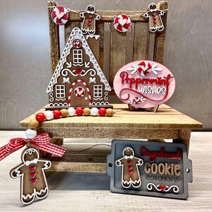 K&ouml;nnte beinhalten: Ein Lebkuchenhaus, ein Lebkuchenmann-Anh&auml;nger und ein Schild mit der Aufschrift "Peppermint Wishes" sind auf einem Holzregal ausgestellt. Ein zweites Schild lautet "Gingerbread Cookie Company".