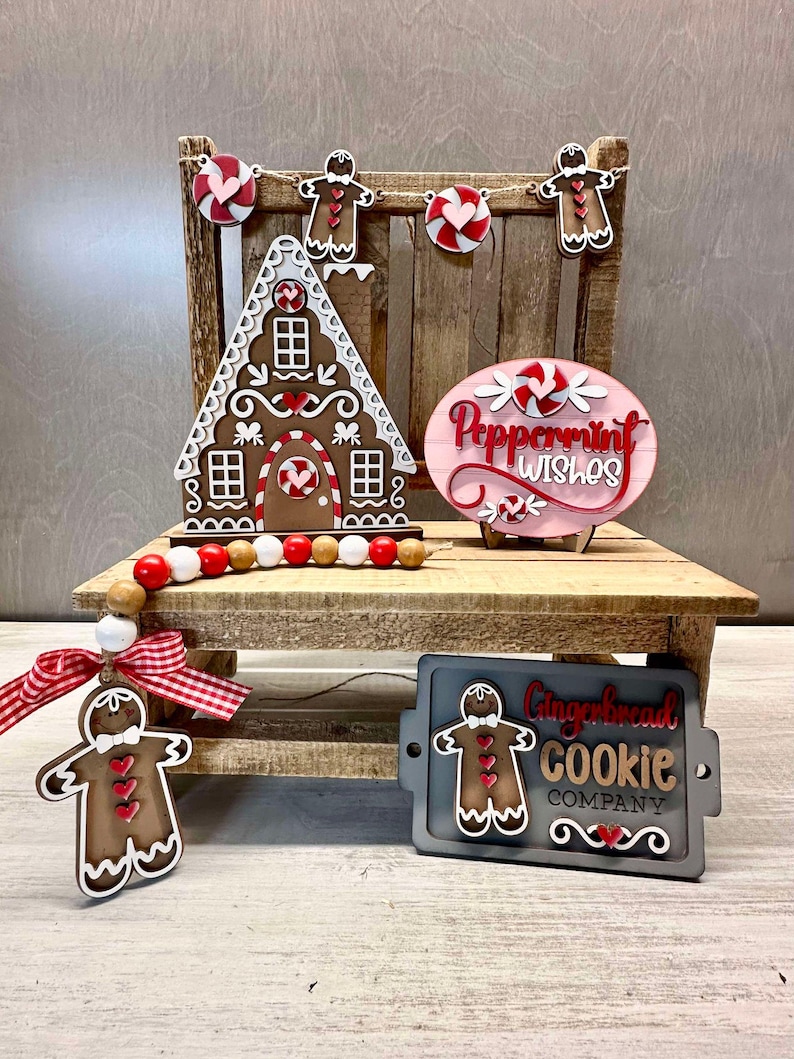 K&ouml;nnte beinhalten: Ein Lebkuchenhaus, ein Lebkuchenmann-Anh&auml;nger und ein Schild mit der Aufschrift "Peppermint Wishes" sind auf einem Holzregal ausgestellt. Ein zweites Schild lautet "Gingerbread Cookie Company".