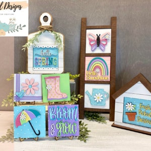 May include: A collection of spring-themed wooden signs with various designs, including a rainbow, a butterfly, a watering can, a daisy, a bird, rainboots, an umbrella, and the words "hello spring", "hello sunshine", "bloom & grow", and "fresh picked daisies".