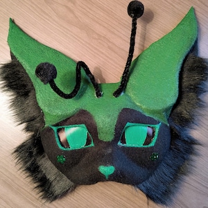 May include: A handcrafted creature mask featuring a black and green felt face with large green mesh eyes, a small green heart-shaped nose, and two black pipe cleaner antennae with pom-poms. The mask is framed by long, dark faux fur.