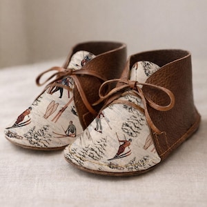 May include: A pair of ankle boots featuring brown textured leather sides and a cream-colored front panel printed with a vintage-style winter scene of people skiing and snowshoeing among pine trees, finished with thin brown leather laces.