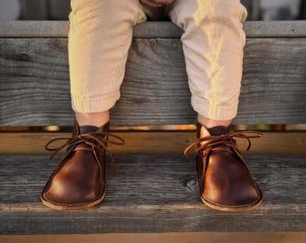 May include: A pair of dark brown leather ankle boots featuring a rounded toe, a low-profile sole, and thin matching brown laces tied in a bow. The boots are constructed with a smooth finish and a simple, clean design that covers the foot.
