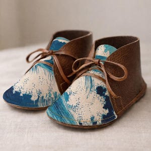 May include: A pair of ankle boots featuring brown textured leather sides and a front panel printed with a blue and cream wave pattern. The boots have thin brown leather laces tied in a bow at the top of the tongue and a low-profile sole.