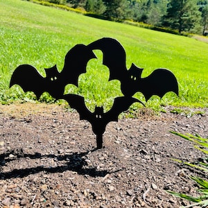 May include: Black metal silhouette of three bats in a circle, with the bats' wings overlapping. The silhouette is on a brown dirt background.