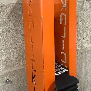 May include: Orange metal wall mounted storage unit with the word "KALLICK" cut out on the front. The unit is holding a stack of black socks.
