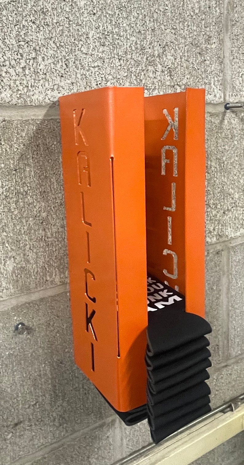 May include: Orange metal wall mounted storage unit with the word "KALLICK" cut out on the front. The unit is holding a stack of black socks.