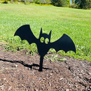May include: Black metal bat silhouette yard decoration. The bat has large eyes and is standing in a bed of brown mulch.