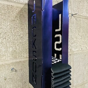 May include: A metal wall-mounted storage rack with the name "JACKSON" cut out on the front. The rack is painted a dark blue color and has a stack of black socks inside.