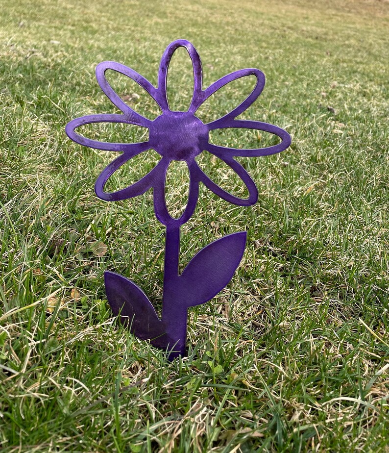 May include: Purple metal daisy flower garden stake. The flower has a simple design with rounded petals and a center circle. The stem has two leaves.