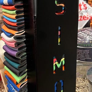 May include: A black metal can holder with the name "SIMON" cut out in the front. The holder is filled with colorful can coolers.