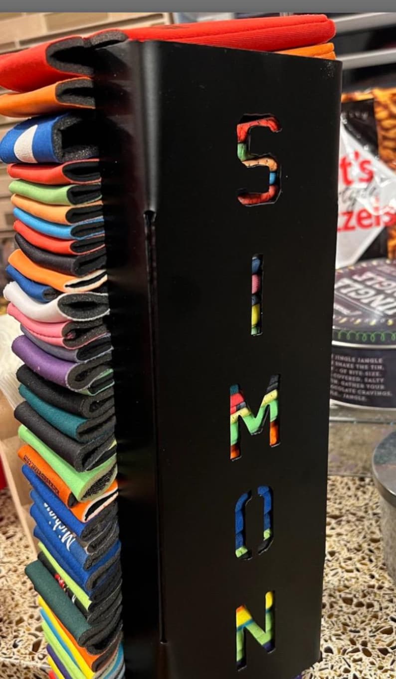 May include: A black metal can holder with the name "SIMON" cut out in the front. The holder is filled with colorful can coolers.