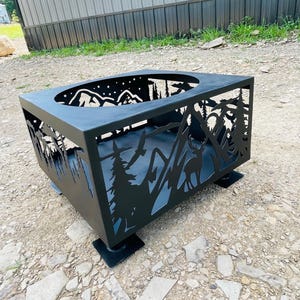 Könnte beinhalten: Schwarzer Metall-Feuerplatz mit einem ausgeschnittenen Design einer Bergszene mit einem Reh, Bäumen und Sternen. Der Feuerplatz ist quadratisch und hat einen erhöhten Rand um die Oberseite.