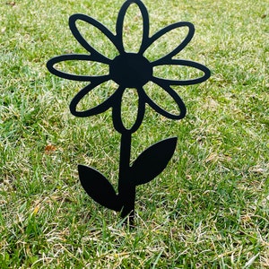 May include: Black metal silhouette of a daisy flower with a stem and two leaves. The flower is stylized with rounded petals and a circular center. The silhouette is standing upright in a grassy area.