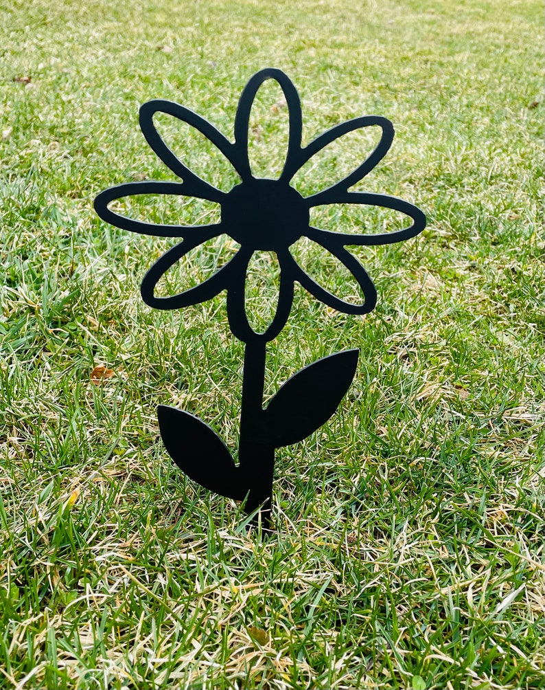 May include: Black metal silhouette of a daisy flower with a stem and two leaves. The flower is stylized with rounded petals and a circular center. The silhouette is standing upright in a grassy area.