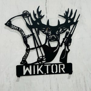 May include: Black metal wall art featuring a deer with antlers, a bow and arrow, and a rifle. The name "WIKTOR" is displayed on a banner below the deer.