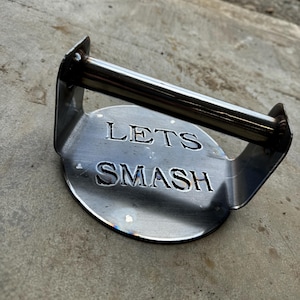 Op de afbeelding: Een zilveren metalen hamburgerpers met een handvat. De pers heeft de woorden "Lets Smash" gegraveerd op de voorkant.