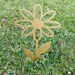 May include: A gold metal daisy flower garden stake. The flower has a simple design with rounded petals and a center circle. The stake is inserted into the ground.