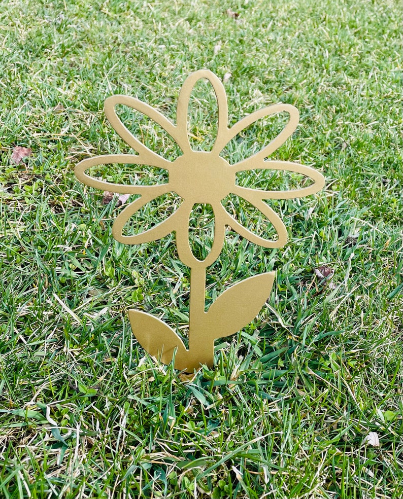May include: A gold metal daisy flower garden stake. The flower has a simple design with rounded petals and a center circle. The stake is inserted into the ground.