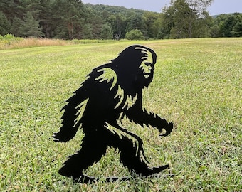 May include: Black metal silhouette of Bigfoot walking in a grassy field. The silhouette is a yard decoration.
