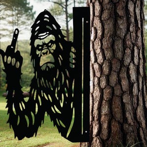 May include: Black metal silhouette of a Bigfoot giving the middle finger, mounted on a tree trunk.