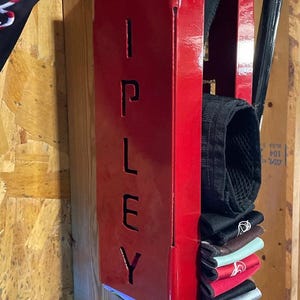 May include: A red metal storage rack with the word "RIPLEY" cut out. The rack is mounted to a wooden wall and holds a stack of black, brown, green, and red fabric wristbands.