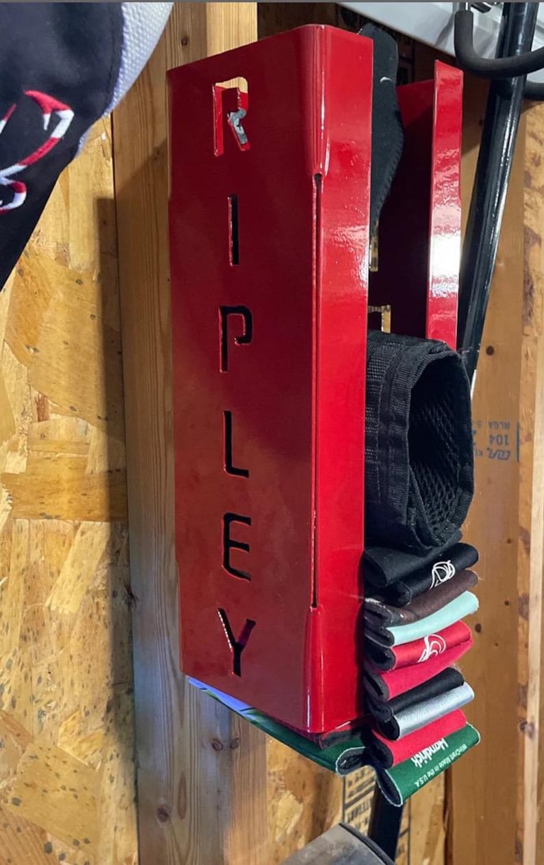 May include: A red metal storage rack with the word "RIPLEY" cut out. The rack is mounted to a wooden wall and holds a stack of black, brown, green, and red fabric wristbands.