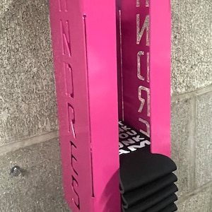 May include: Pink metal wall mounted organizer with the word "PINK" on the right side and "DREES" on the left side. The organizer is holding a stack of black socks.