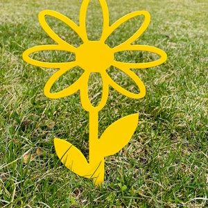 May include: A bright yellow metal daisy flower with a stem and leaves. The flower is standing in a grassy area.