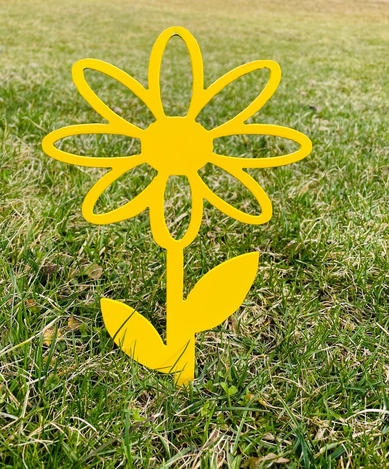May include: A bright yellow metal daisy flower with a stem and leaves. The flower is standing in a grassy area.