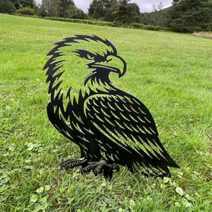 May include: Black metal silhouette of an eagle with detailed feathers and talons. The eagle is standing on a grassy lawn.