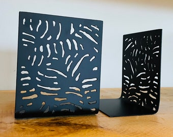 May include: A pair of black metal bookends with a geometric pattern cut-out design. The bookends are on a wooden surface.
