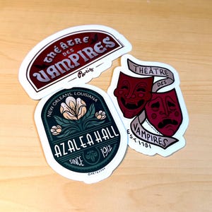 Könnte beinhalten: Drei gestanzte Vinyl-Aufkleber. Einer zeigt einen dunkelroten Bogen mit dem Text Théâtre des Vampires Paris. Ein weiterer ist ein dunkelblaugrünes Oval mit einer Blume und dem Text Azalea Hall New Orleans, Louisiana Since 1912. Der dritte zeigt zwei rote Theatermasken.