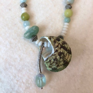 May include: A green and brown seashell pendant necklace with white and green beads. The necklace is made with natural materials and has a rustic, bohemian style.
