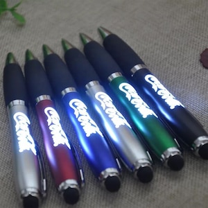 May include: Seven colorful pens with a light-up feature and a stylus tip. Each pen has the word "Coca-Cola" printed on it in white.