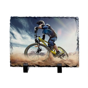 May include: A black slate photo plaque with a printed image of a person riding a yellow mountain bike on a dirt path. The person is wearing a helmet and protective gear. The background is a blue sky with white clouds.