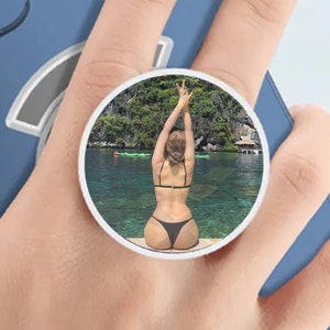 May include: A phone pop socket with a photo of a person in a black bikini standing in a body of water with green water and lush green foliage in the background.