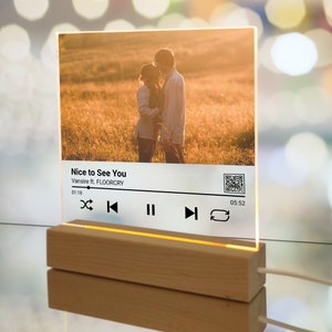 May include: A clear acrylic music player with a wooden base. The player has a photo of a couple in a field, a song title "Nice to See You" by Vanslife ft. FLOORCRY, and a play bar with controls.