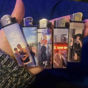 May include: Five personalized lighters with different images printed on them. The lighters are all black with a silver top and bottom. The images include a couple, a woman, a man, and a woman with a neon sign that says "I love you".