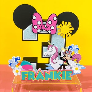 May include: A colorful 3D cake topper featuring a number 3, Mickey Mouse ears, a pink bow, a unicorn, and a Minnie Mouse character riding the unicorn. The topper is on a clear acrylic base and includes the name Frankie.
