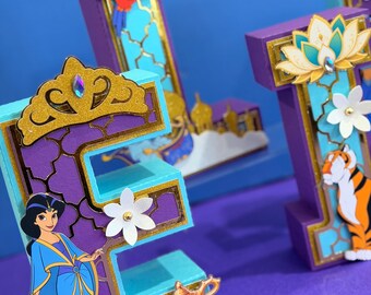Può includere: Una lettera E tridimensionale decorata con una corona glitterata dorata, un fiore bianco e una lampada dorata. Un personaggio dei cartoni animati con un vestito blu è attaccato al lato. La lettera è viola con accenti azzurri e motivi dorati.