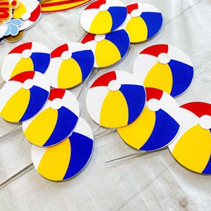 May include: A set of 10 colorful paper beach ball cupcake toppers. Each topper features a white, yellow, blue, and red beach ball design. The toppers are perfect for a summer-themed party.